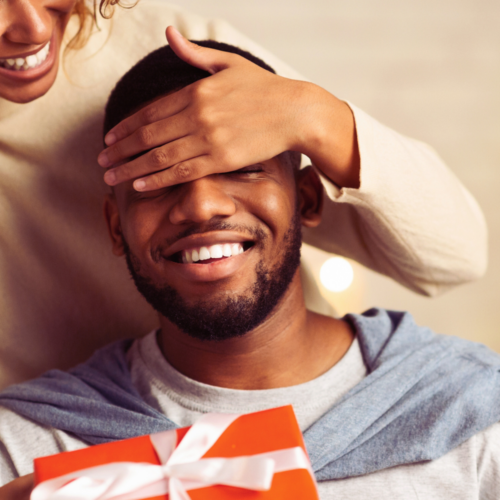 Photo of a girl covering the guys eyes close while handing him a red present. Christmas gift ideas for him