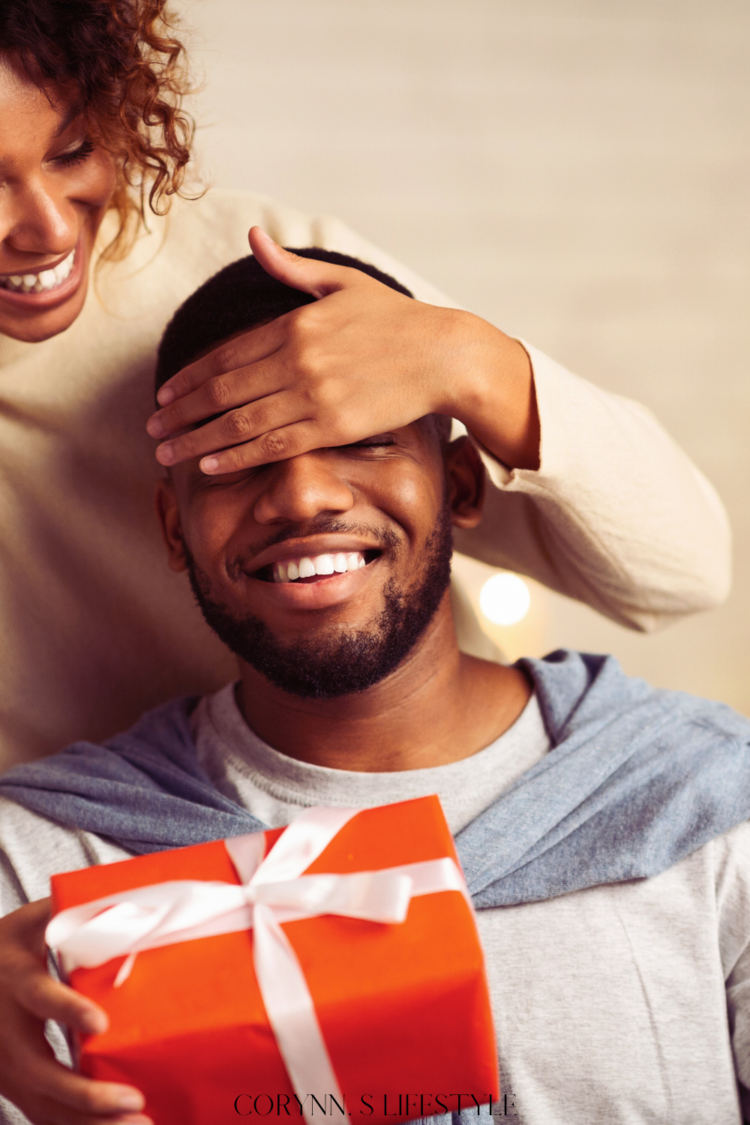 Photo of a girl covering the guys eyes close while handing him a red present. Christmas gift ideas for him